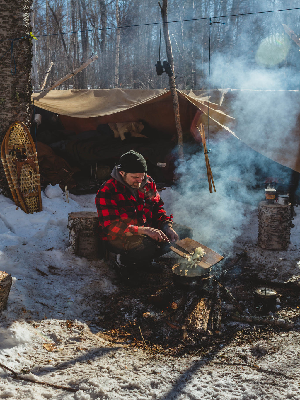Person cooking over a campfire in a snowy forest with a tent and snowshoes in the background.